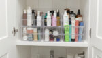 A set of clear, stackable acrylic bins neatly arranged on a bathroom cabinet shelf, filled with skincare and haircare products.